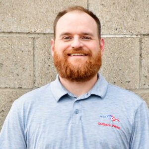 Smiling man with a red beard wearing a light blue polo shirt with an Outback Wrap logo, standing against a concrete block wall.